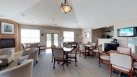 This single-story community clubhouse is thoughtfully designed to serve as a central gathering hub for residents. Upon entry, a prominently positioned reception desk with classic white millwork provides a professional and welcoming focal point. The vaulted ceilings throughout lend an open, airy feel to the entire space, enhanced by an elegant pendant light fixture that anchors the room.

The main lounge area is generously sized and versatile, featuring multiple seating arrangements that accommodate both casual socializing and structured activities. A standout brick fireplace with a traditional mantel serves as a warm focal point on one end of the room, complemented by a wall-mounted flat-screen television and a media console. Comfortable upholstered seating — including a tufted leather sofa, armchairs, and side tables — creates an inviting conversation area nearby.

Multiple dining-height tables with rolling chairs are arranged throughout the space, ideal for card games, activities, or community gatherings. Natural light pours in through numerous large windows and glass-paned French doors that open directly to an outdoor area, providing pleasant views and seamless indoor-outdoor connectivity. Neutral gray carpeting and warm taupe walls contribute to the calm, cohesive aesthetic. A business center and a small refreshment station round out the functional amenities available within this well-appointed community space.