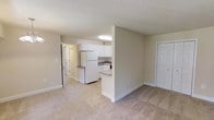 This single-floor space features an open-concept layout that integrates the living area and kitchen into one cohesive environment. The living area is finished with neutral-toned carpeting that runs throughout, contributing to a warm and understated aesthetic. Walls are painted in a soft, light beige tone, creating a clean and airy feel.

The kitchen is partially separated by a half-wall that doubles as a breakfast bar or counter overhang, offering both visual distinction and functional workspace. White cabinetry lines the kitchen walls, providing ample storage, while a white refrigerator and sink are visible within the space. Overhead fluorescent lighting in the kitchen ensures a well-lit cooking area.

A hallway leads from the main living space toward what appears to be additional rooms or a bathroom, with a door visible at the end suggesting a private area such as a bathroom or utility space. A closet with a bi-fold white door is also visible from the living area, offering convenient storage. The overall design is simple, functional, and well-maintained, making the space practical and comfortable.