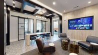 This thoughtfully designed co-working suite spans a single floor and encompasses five distinct functional zones, offering a well-rounded environment for both focused work and collaborative gatherings. The central conference area serves as the heart of the space, anchored by a sleek white communal table surrounded by navy blue chairs with gold-accented legs, beneath a striking coffered ceiling detailed with bold dark beams and a modern linear chandelier. A wall-mounted display screen and natural wood accent wall add a polished, professional touch. Adjacent lounge seating with leather chairs and textured side tables provides a relaxed alternative for informal meetings. Private workspaces include glass-partitioned offices and individual study rooms, each featuring built-in desks, ample natural light from large windows with slatted blinds, and original artwork on the walls. An open workstation area offers a long shared desk with gold-legged task chairs set against a striking three-dimensional mosaic wood tile feature wall, with clerestory windows bringing in additional natural light. A cozy lounge nook with burnt orange chairs and a bold geometric navy and gold accent wall completes the suite's variety of settings. Throughout, curated artwork, a serene landscape mural, rich material contrasts, and sophisticated finishes create an inspiring and cohesive atmosphere.