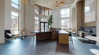 Located at 2600 N Orange Ave in Orlando's vibrant College Park neighborhood, this contemporary single-floor commercial lobby space makes a striking first impression. Soaring double-height ceilings amplify the sense of openness, while expansive floor-to-ceiling windows flood the interior with natural light and offer views of the surrounding urban streetscape. The flooring throughout is finished in large-format dark slate tiles, providing a sleek, grounding contrast to the bright white brick walls.

At the heart of the space sits a substantial central island with a dark stone countertop and warm wood cabinetry, accompanied by bar-height seating — ideal for casual work or conversation. Overhead, an artfully designed pendant light fixture adds a sculptural focal point to the reception area. Lush indoor greenery, including a statement tropical plant, brings life and texture to the interior.

Multiple intimate seating nooks are positioned near the tall windows, each featuring curated accent chairs in teal and neutral tones paired with elegant marble-topped side tables, creating inviting spots for relaxed interaction. A dedicated refreshment bar area is outfitted with a dark stone countertop, mosaic tile backsplash, black matte fixtures, and stainless steel appliances. A living green wall near a glass-enclosed corridor adds a vibrant, biophilic design element, completing this polished and welcoming space.