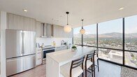 This sophisticated high-rise residence combines contemporary design with breathtaking panoramic views, featuring floor-to-ceiling black-framed windows that frame stunning mountain and cityscape vistas from nearly every room. The open-concept kitchen, dining, and living area serves as the heart of the home, where natural light floods the space throughout the day. The kitchen is outfitted with light wood cabinetry, a white quartz waterfall island with bar seating, stainless steel appliances, a hexagonal tile backsplash, and globe pendant lights that add a refined finishing touch. The living room is anchored by the sweeping corner window views, light hardwood-style flooring, and a clean, airy aesthetic. Both bedrooms offer generous proportions and direct access to the dramatic floor-to-ceiling views, with the spacious primary bedroom providing a serene retreat. The three bathrooms are finished to a high standard, featuring the primary bath with a double-sink vanity in light wood tones, matte black hardware, large-format tile, and a frameless glass walk-in shower. A connecting hallway provides seamless flow between all rooms. The overall design is cohesive, modern, and light-filled, with a neutral palette that highlights the extraordinary outdoor scenery.