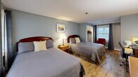 Located at 473 Main St in Barnstable, Massachusetts, this well-appointed guest room offers a comfortable and functional layout across a single floor. The spacious bedroom accommodates two full-sized beds dressed in neutral gray quilted bedding, complemented by warm wood-toned headboards and coordinating nightstands with a classic table lamp. Light hardwood flooring runs throughout, adding warmth to the cool blue-gray painted walls.

Natural light flows in through large windows dressed with dark curtain panels, creating a pleasant and airy atmosphere. A red accent chair provides a cozy reading nook near the window. The room is equipped with a work desk and ergonomic chair, a flat-screen television, a dresser, and a wall-mounted mini-split air conditioning unit for year-round climate control.

A dedicated closet with hanging rod offers convenient storage. The private bathroom features a clean, bright design with white walls, classic red-and-white tile flooring, a combined shower and soaking tub with a curved curtain rod, a single vanity with mirror, and ample towel storage. The overall ambiance is tidy, comfortable, and well-suited for both short-term and extended stays in the heart of Cape Cod.