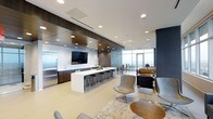 This sophisticated single-floor amenity space showcases a refined, open-plan design that seamlessly blends functionality with contemporary elegance. The layout is anchored by a striking kitchen area featuring dark wood cabinetry, stainless steel appliances, and a long white countertop island lined with sleek bar stools — ideal for casual dining or social gatherings. A wall-mounted flat-screen TV is set against a textured tile backsplash, adding a media-ready focal point to the kitchen zone. Recessed lighting throughout the space is complemented by a bold dark-paneled ceiling detail that adds depth and visual contrast to the otherwise bright, airy interior. The lounge area transitions naturally from the kitchen, offering a curated mix of modern swivel chairs, ottomans, and seating arrangements set atop a layered area rug, creating an inviting space for relaxed conversation or informal meetings. Floor-to-ceiling windows along the far wall flood the interior with abundant natural light and offer open views of the surrounding cityscape, enhancing the sense of spaciousness. The neutral tones of the flooring and walls are thoughtfully offset by warm wood accents and rich upholstered furnishings, resulting in a polished, welcoming ambiance throughout.