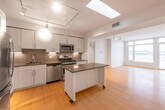 This contemporary single-floor residence offers a thoughtfully designed layout with clean lines and a bright, modern aesthetic throughout. The open-plan kitchen is a standout feature, showcasing white shaker cabinetry, a classic tile backsplash, sleek grey countertops, and a full suite of stainless steel appliances including a gas range, built-in microwave, refrigerator, and dishwasher. A central kitchen island on casters adds flexible prep and dining space, while a skylight overhead floods the area with natural light. Track lighting provides additional illumination throughout the kitchen and living areas, which flow seamlessly together across warm-toned hardwood flooring. Large windows in the bedroom spaces frame urban and outdoor views, allowing abundant natural light to fill each room. The bedrooms are finished with the same hardwood flooring, neutral walls, and clean white trim, creating a calm and versatile environment. The bathrooms are finished with large-format dark slate tile flooring, offering a striking contrast to the light tones found elsewhere. Each bathroom features a vanity with a stone-topped sink, chrome fixtures, and practical storage. The overall ambiance of the space is modern, airy, and move-in ready.