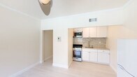 This single-floor space is defined by its clean, bright aesthetic and thoughtfully designed layout. The open living area immediately draws attention to the notably tall ceilings adorned with crown molding, creating a sense of generous vertical space throughout. Light-toned hardwood flooring runs continuously underfoot, contributing to the airy, cohesive feel of the interior.

The kitchen is neatly integrated into the living area and is well-appointed with white shaker-style cabinetry, granite countertops, and a matching granite tile backsplash. Stainless steel appliances include a gas range, over-the-range microwave, and refrigerator, complemented by a stainless steel sink. The kitchen's neutral palette and clean lines align seamlessly with the rest of the space.

The bedrooms are comfortably sized, each featuring light wood flooring, crown molding, and ceiling fans with light fixtures. Windows allow natural light to filter in, with one room offering views of an outdoor pool area through a window with exterior balcony access. The bathroom continues the light, neutral theme, featuring teal tile surrounding a shower-tub combination, a white vanity, and light wood flooring.