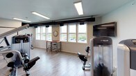 This fitness center is a thoughtfully designed, single-floor facility built to support a well-rounded workout experience. The space features light wood-look flooring throughout, creating a clean and modern aesthetic that is both functional and inviting. Large windows along one wall flood the room with natural light and offer open views of the surrounding outdoor landscape, contributing to an energizing atmosphere.

The facility is outfitted with a comprehensive selection of Precor strength training machines, including a multi-function cable station and a seated leg press, providing users with a variety of resistance training options. Cardio equipment is also present to complement the strength-training offerings. A wall-mounted flat-screen television adds entertainment for those using the cardio machines.

A convenient high-top table with bar stools is positioned near the windows, offering a space to rest or review workout plans. Full-length mirrors on one wall enhance the sense of space and allow users to monitor their form during exercise.

The interior design incorporates bold yellow accent walls contrasted against light gray and white tones, giving the space a dynamic, motivational energy. Inspirational typographic artwork adorns the walls, reinforcing the fitness-focused atmosphere. Recessed panel lighting and exposed industrial ductwork on the ceiling add a contemporary industrial touch to the overall design.