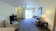 This single-story space centers around a welcoming living room designed for both comfort and functionality. The room is finished with plush wall-to-wall carpeting in a neutral gray-blue tone, providing a soft and cozy underfoot feel throughout the main gathering area. Light-toned walls create a clean, airy atmosphere that complements the natural light streaming in through large sliding glass doors, which open to an outdoor area and serve as a prominent focal point of the room. Vertical blinds on the sliding doors allow for flexible light control and privacy.

A ceiling fan with an integrated light fixture is centrally mounted, offering both overhead illumination and air circulation. The entry door, framed with warm wood trim, is positioned to one side, creating a clear and practical flow into the living space. A dark wood dresser unit near the entry provides additional storage, while framed wall art adds a decorative touch. The room comfortably accommodates ample seating, making it well-suited for relaxing or entertaining. Overall, the space offers a straightforward, functional design with an inviting and comfortable ambiance.