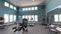 This fitness center is designed to accommodate a comprehensive workout experience within a single, open-plan space. The room's most striking architectural feature is its dramatically tall ceiling, which creates an expansive, airy atmosphere enhanced by dual ceiling fans that promote air circulation throughout. A double row of large windows, including a clerestory bank positioned near the top of the walls, floods the interior with natural light while offering views to the exterior.

The walls are finished in a calming teal-blue tone, complemented by crisp white crown molding and window trim, lending the space a clean, modern aesthetic. The flooring features a herringbone-patterned carpet in a neutral gray tone, adding visual texture underfoot.

Equipment is thoughtfully arranged to maximize usable floor space. Cardio offerings include multiple treadmills and stationary cycling bikes. Strength-training options feature a leg extension/curl machine and a multi-function cable machine suitable for a range of resistance exercises. Wall-mounted flat-screen televisions are positioned for easy viewing during workouts. Recessed lighting supplements the natural light, ensuring the space remains well-lit at all times.
