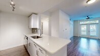 This single-floor unit presents a clean, modern aesthetic defined by neutral tones, wood-style flooring, and an open layout that promotes a sense of spaciousness. The kitchen serves as a central feature, showcasing white cabinetry with sleek hardware, a quartz peninsula with an undermount sink, a tile backsplash, and a full suite of stainless steel appliances including a refrigerator, range, and microwave. Track lighting illuminates the workspace effectively.

The living area is generously proportioned, featuring a ceiling fan and French doors with a transom window above, allowing natural light to pour in and offering views to the outdoors. An adjoining room, suitable as a dining or secondary living space, continues the same flooring and neutral palette.

The bedrooms are well-sized with ceiling fans, ample natural light from large windows with blinds, and open closets with built-in shelving for organized storage. The bathroom is finished with a white quartz vanity, a wood-framed mirror, a tub-shower combination with tile surround, and recessed lighting, maintaining the unit's consistent modern character.

Throughout, bright white walls, clean lines, and cohesive finishes create a polished, move-in-ready environment.