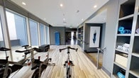 This fitness center offers a thoughtfully designed, single-floor workout space that balances functionality with contemporary style. The interior features light wood-toned flooring that runs the length of the facility, complemented by cool gray walls that create a focused, energizing atmosphere. Recessed lighting throughout ensures the space is well-lit, while large floor-to-ceiling windows along one side invite abundant natural light and provide an open, airy feel.

The cardio zone is prominently featured at the front of the space, housing multiple stationary cycling bikes ideal for high-intensity training sessions. Moving deeper into the facility, the strength training area is visible in the background, equipped with a selection of machines and weight equipment to support a full-body workout regimen. A built-in shelving unit along one wall provides organized storage for fitness accessories and supplies, keeping the floor area clean and uncluttered.

Decorative touches, including a framed feather artwork, add a polished, modern aesthetic that elevates the space beyond a standard gym environment. A storage unit for yoga mats or resistance bands adds further convenience. The overall layout encourages a smooth flow between exercise zones, making this fitness center a well-rounded and visually appealing facility for daily training.