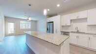 Located at 3018 N Recker Rd in Mesa, AZ, this well-appointed single-story home offers three bedrooms and two bathrooms across a thoughtfully designed floor plan. The open-concept kitchen and living area serves as the heart of the home, anchored by a large center island with granite countertops and pendant lighting overhead. White shaker cabinetry, a subway tile backsplash, and a full suite of stainless steel appliances — including a French door refrigerator, electric range, microwave, and dishwasher — complete the kitchen. Light wood-look flooring flows throughout the main living areas, complemented by neutral warm-toned walls and ceiling fans that enhance comfort. The living room features a ceiling fan, windows for natural light, and direct access to an exterior door. The primary bedroom is a generous retreat with light hardwood flooring, a stylish barn door leading to an ensuite bathroom with granite vanity, walk-in shower, and an adjacent walk-in closet. Secondary bedrooms are carpeted and each include closet storage. The second bathroom mirrors the same high-quality finishes with a granite vanity, white cabinetry, and a shower-tub combination. A hallway and two closets round out the layout, offering practical storage solutions throughout.