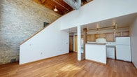 This two-floor loft-style space combines industrial character with warm, functional design. The main level opens into a spacious living area highlighted by soaring ceilings that amplify the sense of openness. A striking exposed brick wall serves as a defining architectural element, complemented by warm wood beam detailing and visible ductwork overhead — hallmarks of authentic loft construction. Rich hardwood flooring runs throughout, adding warmth and continuity across the space.

The kitchen is positioned in an open, semi-enclosed layout with a peninsula counter, warm wood cabinetry, and track lighting overhead, making it both practical and visually connected to the main living area. A staircase with a wood railing leads to the upper level, offering vertical dimension and additional living or sleeping space.

The overall ambiance is industrial yet inviting, balancing raw architectural features — exposed brick, beams, and ductwork — with clean white walls and natural wood tones. The high ceilings and open floor plan create a versatile, airy environment well-suited for a variety of uses. The thoughtful blend of original structural elements and modern finishes gives this space a distinctive character that is both functional and visually compelling.