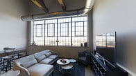 This single-floor space makes a memorable impression with its distinct industrial character and thoughtfully curated design. The living area is anchored by a dramatic wall of large, grid-paned factory-style windows that stretch nearly from floor to ceiling, flooding the interior with abundant natural light and offering sweeping views of the surrounding urban landscape. The generous ceiling height amplifies the sense of openness, while exposed silver ductwork and visible structural beams overhead reinforce the industrial aesthetic with authenticity and purpose. The living space is furnished with a contemporary sensibility — a plush light gray sofa and matching accent chair are arranged around a set of sleek, round white nesting tables atop a deep navy area rug, creating a comfortable and visually cohesive gathering area. A dark, multi-drawer credenza along one wall adds both storage and contrast, complemented by a large flat-screen television. The interplay of raw architectural elements — bare concrete, dark steel window frames, and utilitarian ductwork — against refined furnishings gives the space a dynamic tension that is both stylish and inviting. This space is well-suited for those who appreciate urban loft living with a sophisticated, modern edge.
