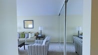 This single-floor space presents a comfortable and functional living area characterized by soft, neutral color tones throughout. The living room is carpeted in a warm beige hue that complements the light sage-green walls, creating a calming and cohesive atmosphere. Seating includes a cushioned sofa adorned with an assortment of decorative pillows in muted greens and grays, alongside a bold houndstooth-patterned accent chair that adds visual interest to the room. A dark wood side table with a classic table lamp provides ambient lighting, while a framed mirror on the wall reflects natural light and contributes to the sense of openness. A notable architectural feature is the sliding mirrored partition, which divides sections of the space while maintaining an airy, open feel. The vaulted ceiling adds a sense of vertical volume to the room, enhancing the overall spaciousness. The neutral palette and understated décor give the space a clean, welcoming ambiance well-suited for relaxed everyday living.