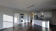 This single-floor unit showcases a contemporary open-concept design that seamlessly connects the living area to the kitchen. The space is finished with rich, dark hardwood flooring that runs throughout, providing a striking contrast against the bright white walls and ceilings. Track lighting spans the ceiling, offering flexible and functional illumination across the main living area.

The kitchen is thoughtfully designed with white shaker-style cabinetry, granite or stone countertops, and a full suite of stainless steel appliances, including a refrigerator, microwave, and range. A peninsula-style island with an undermount sink provides additional workspace and serves as a natural divider between the kitchen and living areas. Pendant lights hang above the island, adding a refined decorative touch.

A dedicated in-unit laundry closet houses a stacked washer and dryer, tucked neatly between two white panel doors. Natural light filters in through windows, brightening the space and highlighting its clean, modern finishes. The overall ambiance is sleek, functional, and well-appointed, making it a highly practical living environment.