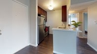Located at 10631 Lindley Ave, this single-floor residence offers a well-organized layout with distinct living, kitchen, and bedroom areas. The living room is a standout space, showcasing vaulted ceilings accented by a rich, dark-stained exposed wood beam that adds warmth and architectural character. Dark hardwood flooring runs throughout the main living areas, complementing the neutral gray walls and clean white baseboards. A ceiling fan with a light fixture provides both comfort and illumination, while a full-length sliding glass door dressed with vertical blinds fills the room with natural light and offers outdoor access. The kitchen is efficiently designed with rich cherry-toned cabinetry reaching toward the ceiling, stainless steel appliances including a refrigerator and range, and a peninsula with a light-colored countertop and built-in sink. The textured peninsula wall adds subtle visual interest. The bedroom features plush wall-to-wall carpeting in a neutral tone, smooth gray walls, and a window with blinds that allows in ample natural light. Overall, the space presents a clean, move-in ready aesthetic with a blend of practical design and inviting architectural details.