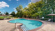 Located at 955 Easton Road in Warrington, Pennsylvania, this outdoor amenity area centers around a generously sized, rounded swimming pool with clear blue water and a classic tiled border. The pool is designated as a no-diving zone and is equipped with safety equipment, including a life ring, ensuring a secure environment for users. Surrounding the pool is a expansive concrete sundeck that provides ample space for relaxation and recreation. The deck is furnished with modern sling-style lounge chairs and several large shade umbrellas, offering comfortable spots to unwind in or out of the sun. The area is enclosed by both an ornamental iron fence and a chain-link perimeter fence, providing defined boundaries while maintaining an open, airy feel. Mature trees line the edges of the property, offering natural shade and a verdant backdrop that enhances the outdoor atmosphere. A small poolside structure, visible in the background, appears to serve as a utility or storage facility. Nearby seating areas with tables and chairs provide additional gathering space. The grounds feature well-maintained green lawns, and the overall setting creates a pleasant, community-oriented outdoor retreat suited for warm-weather enjoyment.