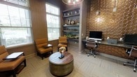 This thoughtfully designed single-floor space combines comfort and functionality within a refined, upscale aesthetic. The room is anchored by a warm color palette of rich browns, golds, and deep taupe tones that create an inviting and sophisticated ambiance throughout.

A cozy lounge area near the window features two cushioned amber-toned armchairs positioned around a distinctive round wooden coffee table with a metallic drum base, ideal for casual conversation or relaxed reading. Large windows with horizontal blinds allow natural light to filter into the space, enhancing its warmth.

A built-in shelving unit with a blue-gray backing serves as a decorative focal point, displaying curated décor including ceramic vases, greenery, and assorted accents that add visual depth and character.

On the opposite side, a functional workstation with a granite-topped desk, ergonomic office chair, and desktop computer is set against a striking textured accent wall featuring a honeycomb-patterned bronze tile, adding a bold architectural element to the space.

Patterned carpeting in a herringbone design grounds the room, tying together its various design elements cohesively.