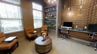 This thoughtfully designed single-floor space combines comfort and functionality within a refined, upscale aesthetic. The room is anchored by a warm color palette of rich browns, golds, and deep taupe tones that create an inviting and sophisticated ambiance throughout.

A cozy lounge area near the window features two cushioned amber-toned armchairs positioned around a distinctive round wooden coffee table with a metallic drum base, ideal for casual conversation or relaxed reading. Large windows with horizontal blinds allow natural light to filter into the space, enhancing its warmth.

A built-in shelving unit with a blue-gray backing serves as a decorative focal point, displaying curated décor including ceramic vases, greenery, and assorted accents that add visual depth and character.

On the opposite side, a functional workstation with a granite-topped desk, ergonomic office chair, and desktop computer is set against a striking textured accent wall featuring a honeycomb-patterned bronze tile, adding a bold architectural element to the space.

Patterned carpeting in a herringbone design grounds the room, tying together its various design elements cohesively.