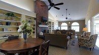 This single-story space is designed to feel open and welcoming, anchored by a dramatic floor-to-ceiling brick fireplace that serves as the room's focal point. A mounted flat-screen television sits above the white mantel, flanked by built-in shelving units with yellow-accented interiors, providing both display and storage space for books and decorative items. The vaulted ceilings add a strong sense of height and volume, enhanced by multiple ceiling fans and pendant lighting that ensure the space remains bright and comfortable. Large arched windows along one wall allow generous natural light to fill the room, contributing to the airy ambiance. The seating area features plush, dark upholstered sofas and armchairs arranged around an area rug, offering a comfortable space for gathering and relaxation. A round wooden dining table with cross-back chairs is positioned in the foreground, providing a dedicated area for casual dining or socializing. The warm, neutral color palette combined with natural textures throughout creates a cozy yet refined atmosphere well-suited for communal use.