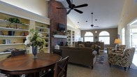 This single-story space is designed to feel open and welcoming, anchored by a dramatic floor-to-ceiling brick fireplace that serves as the room's focal point. A mounted flat-screen television sits above the white mantel, flanked by built-in shelving units with yellow-accented interiors, providing both display and storage space for books and decorative items. The vaulted ceilings add a strong sense of height and volume, enhanced by multiple ceiling fans and pendant lighting that ensure the space remains bright and comfortable. Large arched windows along one wall allow generous natural light to fill the room, contributing to the airy ambiance. The seating area features plush, dark upholstered sofas and armchairs arranged around an area rug, offering a comfortable space for gathering and relaxation. A round wooden dining table with cross-back chairs is positioned in the foreground, providing a dedicated area for casual dining or socializing. The warm, neutral color palette combined with natural textures throughout creates a cozy yet refined atmosphere well-suited for communal use.