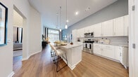 This single-floor residence is defined by its open-concept layout and striking soaring ceilings that create an impressive sense of volume throughout the main living area. The kitchen is a standout feature, outfitted with crisp white shaker cabinetry, granite countertops, a full suite of stainless steel appliances — including a range, microwave, dishwasher, and refrigerator — and a large center island with bar seating, illuminated by elegant pendant lighting. The kitchen flows seamlessly into the dining and living areas, where warm wood-look flooring and floor-to-ceiling windows flood the space with natural light and provide views to the outdoor surroundings. The living room offers a relaxed yet sophisticated atmosphere with ample space for comfortable seating arrangements. The primary bedroom is a serene retreat, featuring plush carpeting, a bold accent wall, tall ceilings, and large windows that invite natural light. It connects directly to a spacious walk-in closet equipped with multi-tier wire shelving and to an en-suite bathroom showcasing a double-sink granite vanity and wood-look flooring. The overall palette of soft blues, warm whites, and natural wood tones lends the space a cohesive, modern aesthetic throughout.