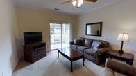 This single-floor residence offers a welcoming and functional layout designed for comfortable everyday living. The living room serves as the heart of the space, featuring soft carpeted flooring and warm, neutral-toned walls that create a relaxed and inviting atmosphere. A ceiling fan with an integrated light fixture is centrally positioned overhead, providing both airflow and ambient lighting throughout the room. A large sliding glass door fills the space with natural light and opens directly onto a covered balcony, offering a pleasant outdoor retreat with views of surrounding greenery. The living room is well-appointed with a substantial leather sofa, a classic wooden coffee table with elegant cabriole legs, and a dark wood media console that houses a flat-screen television. A framed mirror on the wall adds depth and visual interest to the room. Crown molding details along the ceiling contribute a polished, refined touch to the overall design. The seamless connection between the interior living space and the outdoor balcony enhances the sense of openness, making this a practical and comfortable home suitable for a variety of lifestyles.