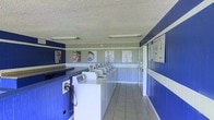 This single-story laundromat is designed with a practical and functional layout, offering a straightforward self-service experience. The interior is immediately distinguished by its bold blue and white color scheme, with blue paneled walls accented by white horizontal trim that runs along the perimeter, creating a clean and structured visual identity.

The main area is lined with a row of top-loading washing machines arranged in an L-shaped configuration, maximizing the use of available floor space. The machines are neatly aligned along the walls, providing ample capacity for multiple users at once. A built-in counter runs along one side, offering a convenient surface for sorting and folding laundry.

The flooring consists of light-colored square tiles that are easy to maintain and contribute to the room's clean appearance. Overhead, fluorescent light fixtures mounted to the textured ceiling provide bright, even illumination throughout the space. Informational and instructional posters are displayed on the walls, offering guidance to users. The overall ambiance is utilitarian, well-organized, and clearly designed to prioritize efficiency and ease of use.