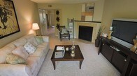 This single-floor residence offers a thoughtfully designed layout centered around a welcoming living room. The space is finished in warm, neutral tones with light beige walls and plush carpeted flooring throughout, creating a cozy and inviting atmosphere.

The living room serves as the focal point of the home, anchored by a tile-surround fireplace with a classic mantle that adds both warmth and architectural character. A large mirror above the mantle enhances the sense of openness in the room. The space comfortably accommodates a full sofa, accent chair, and a central coffee table, making it well-suited for both relaxing and entertaining.

A tall bookcase and decorative wall accents add personality to the room, while a table lamp provides soft ambient lighting. The living area flows naturally into a hallway that connects to the remaining rooms of the residence, suggesting a practical and cohesive floor plan. Closet doors are visible along the hallway, indicating ample built-in storage throughout.

The overall ambiance of the space is calm and neutral, with a traditional aesthetic supported by dark wood furniture accents and understated décor. Natural and artificial lighting work together to keep the interior feeling bright and comfortable. The layout is efficient, making strong use of the available space on a single level.