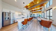 This single-floor open-concept space showcases a striking architectural design defined by soaring vaulted ceilings with exposed warm-toned wood trusses and steel beams, creating a dramatic and airy atmosphere throughout. Light hardwood flooring flows seamlessly across the entire space, unifying the kitchen, dining, and living areas.

The kitchen is thoughtfully designed with crisp white cabinetry, a white subway tile backsplash, stainless steel appliances including a French door refrigerator, a professional-grade range with wall-mounted hood, and a large center island with quartz countertops and leather bar seating. Pendant lighting with textured glass globes adds a refined touch overhead.

The dining area accommodates a generously sized rustic wood table with cross-back chairs and tufted leather seating, positioned directly in front of an expansive wall of black-framed windows that frame outdoor views of a pool and lush greenery, flooding the space with natural light.

The living room features a bold floor-to-ceiling stone tile accent wall housing a built-in fireplace and wall-mounted television flanked by floating wood shelves. Large black-framed windows continue the outdoor connection. The overall ambiance balances rustic warmth with contemporary sophistication, making it a highly functional and visually impressive communal space.