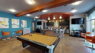 This energetic single-floor community lounge is thoughtfully designed for social gatherings and recreational fun. The open-plan layout is anchored by a full-size pool table at its center, complemented by a shuffleboard table positioned near floor-to-ceiling windows that flood the space with natural light and offer outdoor views of the surrounding landscape. Vibrant teal and orange accent walls, paired with polished concrete floors, create a lively and contemporary atmosphere throughout.

A curved bar area with a mosaic tile backsplash serves as the social hub of the room, featuring bar-height seating, pendant lighting, and under-counter appliances. Adjacent to the bar, a well-appointed kitchenette offers dark cabinetry, granite-style countertops, a wine cooler, stainless steel refrigerator, and a coffee station — ideal for hosting events. A large-format Connect Four game adds a playful touch to the lounge area, which also includes stylish orange accent chairs and a glass bistro table. Bold abstract artwork adorns the walls throughout, enhancing the modern, creative character of the space. Multiple flat-screen televisions are strategically placed for added entertainment, making this clubroom a dynamic and inviting destination for leisure and community connection.
