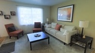 This single-floor space features a living room designed for comfort and functionality. The room is finished with wall-to-wall carpeting and painted in warm, neutral tones that create a welcoming atmosphere. A large window along one wall allows an abundance of natural light to fill the space, offering views of surrounding greenery outside.

The living room is centered around a spacious cream-colored sofa accented with terracotta throw pillows, complemented by a rust-toned accent chair and a matching ottoman. A dark-framed rectangular coffee table anchors the seating arrangement, providing a practical focal point for the room. A framed landscape artwork with warm orange and green hues adorns the main wall, adding a decorative element that ties the color palette together.

A floor lamp positioned near the accent chair provides additional ambient lighting. The overall layout is thoughtfully arranged to maximize the sense of space, making the room feel open and livable. The combination of soft textiles, neutral wall colors, and natural light contributes to a relaxed and inviting ambiance throughout the space.