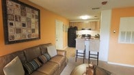 This single-floor space offers an open and connected layout where the living area and kitchen share a cohesive, flowing design. The living room is warm and inviting, anchored by rich warm-toned walls that create an energetic yet comfortable ambiance. The space accommodates comfortable seating and is finished with light tile flooring that transitions throughout the common areas.

The kitchen is efficiently designed and positioned at the heart of the unit, featuring light wood cabinetry that provides ample storage, a black refrigerator, and a gas range. A breakfast bar with tall barstool seating separates the kitchen from the living area, creating a natural social hub ideal for casual dining or conversation. The kitchen's layout maximizes functionality while maintaining an open feel with the adjoining living space.

A textured ceiling adds subtle architectural character throughout, while recessed and flush-mount lighting ensures the space remains well-lit. A private entry door is visible from the living area, and HVAC vents are integrated discreetly into the ceiling. The overall design of this single-floor space balances practicality with a welcoming atmosphere, making it a functional and comfortable place to call home.