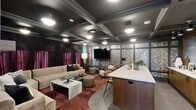 Located at 2902 N Arthur Ashe Blvd in Richmond, VA, this single-floor amenity space offers a compelling blend of sophisticated design and functional versatility across three distinct areas. The centerpiece is a dramatic lounge and bar area distinguished by a coffered, dark-painted ceiling with elegant crown molding and flush-mounted decorative fixtures. Rich burgundy carpeting, velvet drapes in deep plum, and textured metallic wallcoverings create a moody, upscale atmosphere. A generously sized white quartz island with warm wood cabinetry serves as the bar's focal point, surrounded by leather-and-metal bar stools, while the lounge area offers plush neutral sectional seating, a marble coffee table, and a wall-mounted flat-screen TV. A long corridor connects to a bright reading lounge bathed in natural light from floor-to-ceiling windows. Built-in wood bookshelves line the walls, filled with curated volumes and decorative objects, lending a library-like warmth. This area opens to an intimate seating nook furnished with a salmon-toned sofa, patterned rug, and lush greenery framed by dark wood columns. A smaller private room features a striking black wallpaper with a repeating illustrative motif, adding a whimsical yet refined finishing touch to this thoughtfully designed space.