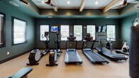 Located at 1951 Old Haywood Rd in Asheville, NC, this fitness center is a thoughtfully designed single-floor facility that balances aesthetic appeal with comprehensive workout functionality. The space is immediately distinguished by its striking architectural details, including exposed natural wood beam ceilings with recessed lighting and ceiling fans that contribute both to the visual warmth of the room and to air circulation during workouts.

The walls are finished in a bold teal color that creates an energizing atmosphere throughout. Large floor-to-ceiling mirrors line the walls, visually expanding the space while allowing users to monitor their form. Multiple windows with blinds admit natural light, offering views of the surrounding greenery.

The cardio zone is equipped with treadmills, elliptical trainers, and stationary bikes, each accompanied by mounted flat-screen televisions for entertainment. The strength training area features a power rack, cable machines, adjustable benches, and a full dumbbell rack with battle ropes and plyo boxes. The entry area includes a wood-accented storage unit, waste receptacle, and glass double doors leading to an adjacent space. Light-toned flooring ties the entire facility together, creating a clean, functional, and welcoming environment.