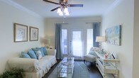 This single-floor living room exudes a light and airy coastal aesthetic, thoughtfully designed with a neutral palette accented by soft shades of blue. The room centers around a white slipcovered sofa adorned with blue and floral throw pillows, complemented by an elegant tufted accent chair in cream. A mirrored glass coffee table anchors the seating arrangement, reflecting light throughout the space and adding a touch of glamour.

Dark hardwood flooring provides a rich contrast to the pale walls, while a soft area rug in muted tones grounds the seating area. Framed wall art and table lamps on either side of the room contribute to its balanced, symmetrical layout.

Natural light flows generously into the space through a set of French doors flanked by two windows, all dressed with white horizontal blinds and floor-length blue draping curtains that enhance the coastal theme. A classic ceiling fan with an integrated light fixture is centrally positioned overhead, ensuring both comfort and illumination. The overall ambiance is serene, welcoming, and elegantly casual.