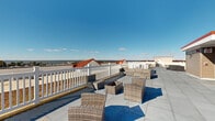 Located in Stevensville, MD, this rooftop terrace offers a generous open-air retreat with sweeping panoramic views of the surrounding Chesapeake Bay region and its natural landscape. The terrace is finished with large-format, smooth concrete pavers that extend across the entire rooftop, providing a clean, modern surface that defines the space.

Multiple seating arrangements are distributed thoughtfully across the rooftop, featuring wicker and rattan lounge chairs, sofas, and a central fire pit table, creating distinct zones ideal for relaxing or entertaining. A classic white picket-style perimeter railing runs the length of the terrace, providing both safety and an unobstructed sightline to the open sky and water views beyond.

A rooftop structure clad in neutral lap siding with a red-accented roofline serves as a stairwell access point and provides a visual anchor to the space. Stone-accented columns add an architectural detail that complements the overall design. Deck storage boxes are conveniently tucked near the structure, offering practical storage without interrupting the open feel of the terrace.

The rooftop's expansive footprint allows for ample circulation space between seating areas. Bathed in natural sunlight from all directions, this outdoor space delivers a bright, airy ambiance with far-reaching views that make it an exceptional venue for outdoor living above the rooftops of Stevensville.