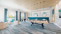 Located in Panama City Beach, Florida, this single-floor recreational lounge space delivers a cohesive coastal aesthetic through a carefully curated combination of design elements and furnishings. The room's interior is anchored by a striking abstract carpet in shades of teal, blue, and grey, evoking the look of weathered driftwood or shallow ocean waters. A warm wood-paneled ceiling with recessed lighting and pendant fixtures adds architectural depth and a natural counterpoint to the crisp white walls.

The space is designed for social activity, prominently featuring a light wood pool table with angular X-frame legs and a full-size foosball table. Along one wall, a continuous teal velvet banquette hosts a row of round-pedestal café tables paired with sleek black chairs, each accented by circular gold-framed mirrors and sculptural teal glass pendant lights above. A chevron-patterned accent wall serves as a backdrop for a wall-mounted television nearby.

Large windows dressed in teal draping curtains allow natural light to fill the space and offer outdoor views of what appears to be a pool area with palm trees. Additional lounge seating with teal and neutral upholstery is positioned near the windows. Ocean-inspired artwork punctuates the walls, reinforcing the refined, beachy ambiance throughout.