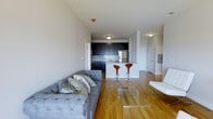 This single-floor residence offers a thoughtfully designed open-concept layout that seamlessly connects the living and kitchen areas. The living room is anchored by a tufted gray sofa and a sleek glass coffee table, creating a comfortable and contemporary gathering space. Warm hardwood flooring runs throughout, lending a sense of continuity and natural warmth to the interior.

The kitchen is a standout feature, showcasing dark navy cabinetry that contrasts beautifully with a classic white subway tile backsplash and stainless steel appliances. A white peninsula with striking red bar stools serves as both a functional prep area and a casual dining spot, adding a bold pop of color to the space.

The open corridor leading away from the main living area hints at additional rooms, suggesting a well-organized floor plan that balances communal and private spaces. Neutral-toned walls and flush-mount ceiling lighting keep the atmosphere bright and airy. The overall design blends modern aesthetics with practical living, making it a well-appointed and inviting space suited for everyday comfort and entertaining alike.