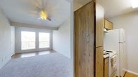This single-floor residence offers a practical and comfortable layout designed to maximize everyday living. The living area is finished with soft wall-to-wall carpeting, creating a warm and inviting atmosphere, while a ceiling fan overhead provides year-round comfort and adds a classic touch to the space. Two windows positioned side by side allow natural light to fill the room, contributing to an open and airy feel. Adjacent to the living area, a tiled entryway or transitional zone provides a durable, easy-to-clean surface near the access point to the space. The kitchen is situated just off the main living area, separated by a distinctive full-height wooden panel that adds a natural, earthy element to the interior. The kitchen features warm-toned wood cabinetry that runs along both upper and lower sections, offering ample storage. A white range and refrigerator complete the functional cooking space, providing all the essential appliances needed for daily meal preparation. The overall design of the space reflects a straightforward, efficient use of the floor plan, with a neutral color palette throughout that lends itself well to a variety of décor styles. The combination of carpet, tile, and natural wood tones gives the interior a grounded, cohesive character.