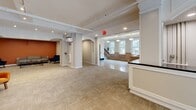 Located at 6 Chatham St in Worcester, Massachusetts, this thoughtfully designed single-floor space offers a generous and versatile layout suited for professional or community use. The welcoming entry hallway immediately impresses with its polished terrazzo tile flooring, elegant pendant lighting, and classic double French doors that allow natural light to pour in from the street. Wainscoting and crown molding details lend a refined, traditional character to the entryway, which also features a reception counter and staircase access. Decorative arched openings connect the hallway to the broader interior, creating a sense of flow and openness throughout. The living area transitions seamlessly from the entry, offering carpeted flooring, recessed lighting, and a bright, open layout. A bold terracotta accent wall anchors one seating lounge, furnished with a contemporary gray sofa and navy accent chairs. A secondary lounge features vibrant orange velvet chairs paired with a glass-top coffee table beneath striking white arched architectural details and neutral gray walls. The space as a whole balances classic architectural elements — including exposed beam ceilings, arched passageways, and detailed millwork — with modern furnishings and a warm, inviting color palette that makes it both functional and visually engaging.