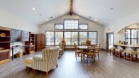 This single-floor community facility is thoughtfully designed to provide residents with a variety of shared spaces that balance comfort and functionality. The welcoming entryway features soaring vaulted ceilings, arched architectural details, warm wood-look flooring, and graceful archways that lead deeper into the building, setting an inviting tone throughout.

The main lounge area is a standout space, boasting dramatic vaulted ceilings accented by an exposed wood beam and an expansive wall of large, grid-paned windows that flood the room with natural light and offer outdoor views. Comfortable upholstered seating, a dining table, and a media unit with a flat-screen television make this an ideal space for relaxing or socializing.

Adjacent to the lounge, the kitchen is accessed through an elegant arched opening and features gray cabinetry, tile backsplash, a full-size refrigerator, pendant lighting, and a peninsula counter with bar seating — well-suited for communal gatherings and casual meals.

The multipurpose activity room offers generous table space with modern seating and built-in cabinetry with a utility sink, providing a versatile environment for classes, events, or crafts. The dedicated computer lab is equipped with multiple workstations, printers, conference-style tables, and colorful seating, making it a practical resource hub for residents.