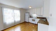 This single-floor space offers a clean, functional layout designed for comfortable everyday living. The kitchen is a standout feature, outfitted with crisp white cabinetry that extends to the ceiling for ample storage, dark granite-style countertops, and a full suite of white appliances including a refrigerator, electric range, and a sink positioned beneath upper cabinets. The warm-toned hardwood flooring runs throughout the space, adding a sense of continuity and warmth to the interior. Natural light plays a key role in the ambiance of this space — a large sliding glass door framed by vertical blinds floods the area with bright, diffused daylight, visually expanding the space and creating an open, airy feel. The transition between the kitchen and the adjacent living area is seamless, reinforced by the consistent flooring and neutral wall tones that keep the space feeling cohesive and inviting. Overhead, a central flush-mount light fixture provides additional illumination, ensuring the space remains well-lit at all hours. The overall aesthetic is understated and practical, with a neutral palette that offers a versatile backdrop for a variety of interior styles.