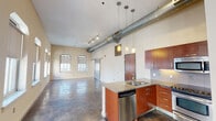 This single-floor urban loft showcases an industrial-modern aesthetic throughout, blending raw architectural elements with refined finishes. The open-plan living and kitchen area commands attention with soaring ceilings, exposed silver ductwork running the length of the space, and polished concrete floors that reflect natural light throughout. A row of arched windows lines the main living area, offering striking views of the surrounding cityscape while bathing the interior in abundant daylight.

The kitchen is a functional centerpiece, featuring rich warm-toned cabinetry, granite countertops on both the island and perimeter surfaces, a stainless steel appliance suite including a refrigerator, range, and microwave, and pendant lighting suspended from the high ceiling. The kitchen island includes a double sink and a built-in dishwasher, providing ample workspace and storage.

The bedroom areas maintain the same polished concrete flooring and tall ceilings, with additional arched windows continuing the theme of natural light and urban views. A sliding barn-style door adds a modern industrial touch between spaces. The bathroom is finished with neutral tile surrounding a full soaking tub and shower combination, complemented by granite vanity countertops and warm wood cabinetry, creating a clean and cohesive look throughout the unit.