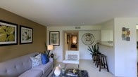 This single-floor residence offers a thoughtfully designed living area that balances comfort and style. The main living room is anchored by a plush sofa adorned with decorative blue and patterned throw pillows, complemented by a glass-topped coffee table resting on a textured area rug. A warm tan accent wall provides a rich contrast to the surrounding white walls, while framed artwork adds visual interest throughout the space.

A circular metallic wall sculpture serves as a standout decorative element, drawing the eye and adding an artistic focal point to the room. Tall indoor plants introduce a natural, organic touch, enhancing the overall ambiance. A white accent chair with a patterned cushion provides additional seating, contributing to the room's layered, inviting feel.

To one side, a counter-height bar area with dark wood stools and white cabinetry above suggests a convenient casual dining or entertaining space. A central hallway visible from the living area offers a glimpse into a softly lit bedroom beyond, hinting at a well-connected and functional floor plan throughout the home.