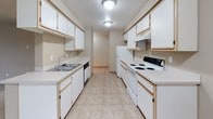 This single-floor residence offers a practical and comfortable layout designed for everyday living. The open living area is generously sized, featuring neutral-toned carpet, a wall-mounted fireplace, and a sliding glass door that fills the space with natural light and provides access to the outdoors. The galley-style kitchen is efficiently organized, with ample white cabinetry lining both walls, light countertops, a double stainless steel sink, a dishwasher, and a full suite of white appliances including a range and refrigerator. Tile flooring runs throughout the kitchen for easy maintenance. The space includes two carpeted bedrooms, each finished with neutral walls and windows that invite natural light. One bedroom features a sliding-door closet, while another offers open access to adjoining spaces. Two bathrooms serve the home: one full bathroom with a soaking tub, a single vanity, and tile flooring, and a second bathroom featuring a walk-in glass shower, an extended vanity with generous counter space, a large mirror, and warm lighting. Throughout the home, textured ceilings and a consistent neutral color palette create a cohesive, understated aesthetic suited to a variety of furnishing styles.