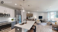 This single-floor residence showcases a thoughtfully designed open-concept layout where the kitchen, dining, and living areas flow seamlessly together. The kitchen is a standout feature, equipped with stainless steel appliances, a two-tone cabinet scheme pairing white uppers with gray lowers, a white subway tile backsplash, and a generously sized quartz-topped island with bar seating. Glass pendant lights hang overhead, complementing the light wood-tone flooring that extends throughout the space. The living area offers a relaxed, airy ambiance with neutral tones and ample room for entertaining.

The bedroom provides a tranquil retreat with large floor-to-ceiling windows and direct access to a private balcony, filling the room with natural light and outdoor views. A dedicated walk-in closet with wire shelving systems offers practical storage. A flex room with expansive windows and balcony access serves well as a home office or secondary lounge.

The bathrooms are finished with attention to detail, including a glass-enclosed walk-in shower with herringbone tile, gray vanity cabinetry, and modern fixtures. Light wood flooring and recessed lighting carry the cohesive, contemporary aesthetic throughout the entire space.