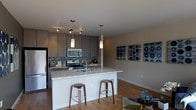 This single-floor space showcases a thoughtfully designed open-concept layout that seamlessly connects the kitchen and living area. The kitchen is a standout feature, equipped with stainless steel appliances including a range, microwave, and refrigerator. Rich granite countertops extend across a spacious kitchen island, which doubles as a breakfast bar with stylish wood bar stools—perfect for casual dining or entertaining. Taupe flat-panel cabinetry with sleek hardware lines the walls, complemented by a mosaic tile backsplash that adds texture and visual interest. Pendant lighting hangs above the island, while a track lighting system over the cooking area ensures excellent task illumination. The warm hardwood flooring flows throughout, tying the kitchen and living spaces together cohesively. Neutral-toned walls provide a clean backdrop, enhanced by contemporary wall art that adds a touch of personality to the open living area. The overall ambiance is modern and inviting, with a well-balanced combination of functional design and stylish finishes that make this space both practical and visually appealing.