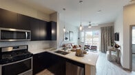 This single-floor residence showcases a contemporary design with a cohesive, modern aesthetic throughout. The open-concept kitchen and living area form the heart of the space, where bold dark cabinetry with flat-panel doors contrasts sharply against white quartz countertops and a white subway tile backsplash. Stainless steel appliances, including a gas range, microwave, and French door refrigerator, complete the well-appointed kitchen. A peninsula with pendant lighting provides additional prep space and casual seating, flowing naturally into the living area.

The living room benefits from abundant natural light through large sliding glass doors that lead to a private balcony, offering outdoor views of the surrounding urban landscape. The bedroom features carpet flooring, dual windows with city views, and a calm, neutral color palette. The bathroom is finished with wood-look flooring, a dark vanity topped with white counters, and a backlit mirror, with direct access to a walk-in closet offering built-in shelving and hanging space. Light wood-look flooring runs throughout the main living areas, tying the space together with a warm, modern finish.
