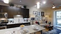 This single-floor space showcases an open-concept layout that seamlessly connects the kitchen, dining, and living areas into one cohesive environment. The kitchen is outfitted with rich dark-brown cabinetry paired with a light stone tile backsplash, creating a striking contrast against the neutral walls. A stainless steel sink and under-cabinet appliances contribute to the kitchen's functional and modern aesthetic, while pendant lighting adds warmth overhead.

The dining area features a white marble-style table with place settings, positioned centrally to bridge the kitchen and living space. The living area is anchored by a neutral-toned sofa accented with bold purple and patterned throw pillows, adding a pop of color. A curated gallery wall displaying an eclectic mix of black-and-white framed artwork serves as a focal point, adding depth and personality to the space.

Recessed lighting throughout, complemented by floor lamps and pendant fixtures, creates a layered lighting scheme. The overall ambiance is sophisticated yet inviting, with a careful balance of dark and light tones, decorative greenery, and stylish accents throughout.
